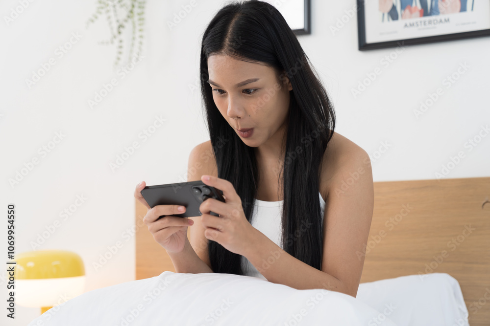 Cheerful young Asian woman playing games on mobile phone on bed in the bedroom Cheerful young Asian woman playing games on mobile phone on bed in the bedroom