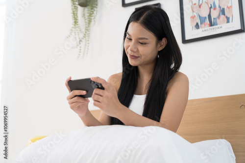 Cheerful young Asian woman playing games on mobile phone on bed in the bedroom