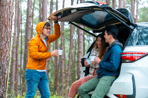 Asian man and woman sitting in car trunk and drinking coffee during road trip travel at natural park. People enjoy outdoor active lifestyle hiking and camping at forest mountain on holiday vacation.