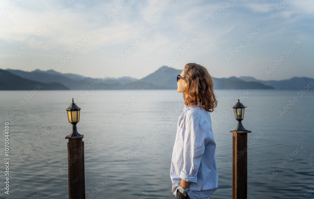 Fototapeta premium Young woman standing on the pier in the morning admiring the sea and mountain landscape