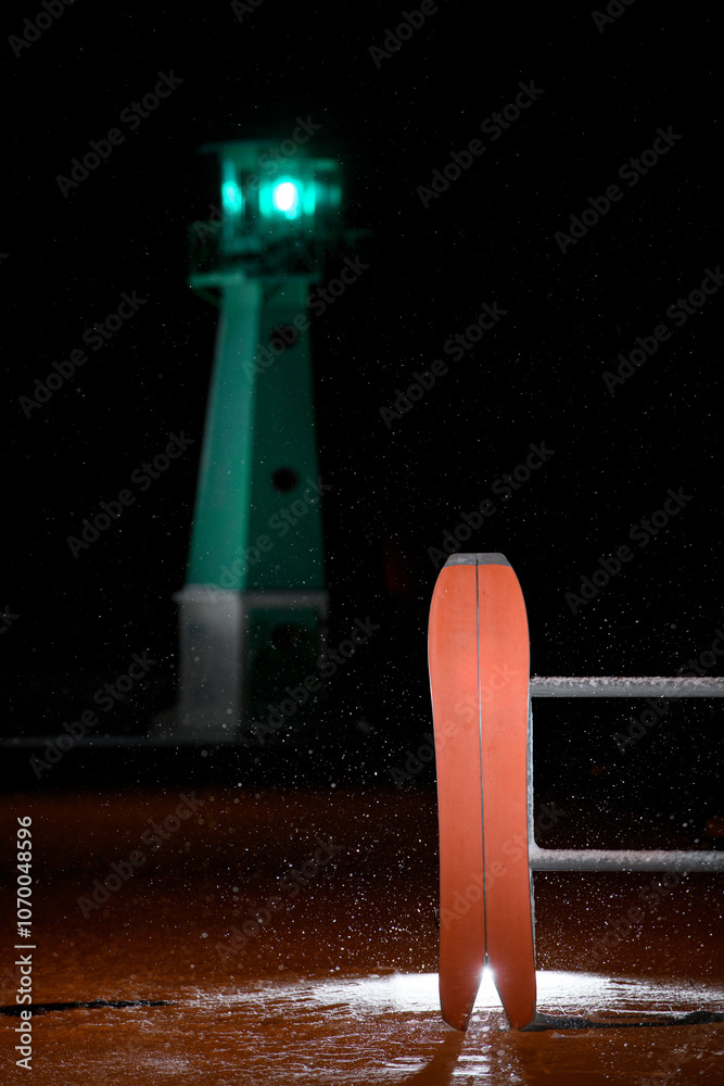 Fototapeta premium Red splitboard is standing, leaning against a fence, on the sand against a background of a glowing pendulum