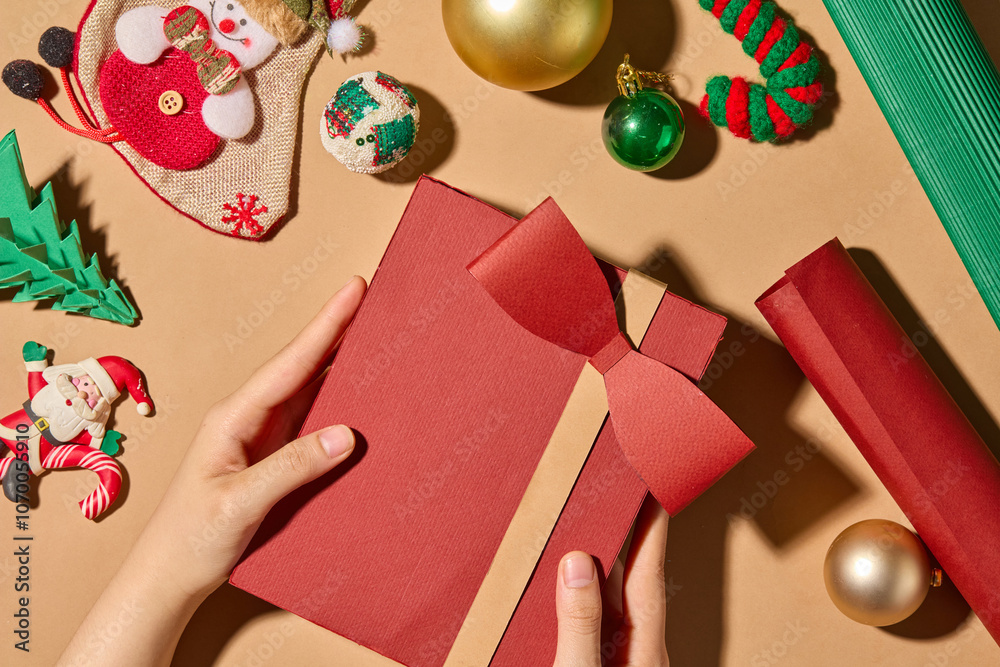 Fototapeta premium Top-down close-up photo Christmas theme showing a hand holding a red gift box. On a brown surface are Christmas themed decorations such as candy cane, Christmas balls, paper pines, and rolls of paper.