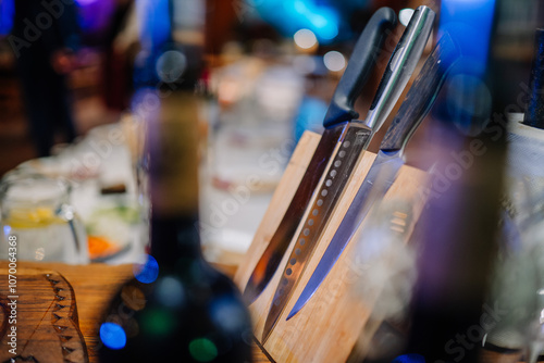 Fotografija A set of kitchen knives with various blades is displayed in a wooden knife block on a table, surrounded by blurred background elements and lights