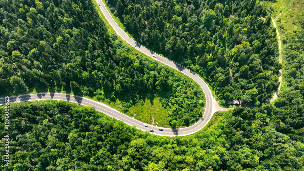 Winding mountain road through dense green forest. Aerial view of a winding road cutting through a lush, dense forest in a mountainous area, with cars navigating the curves. Winding mountain road through dense green forest. Aerial view of a winding road cutting through a lush, dense forest in a mountainous area, with cars navigating the curves.