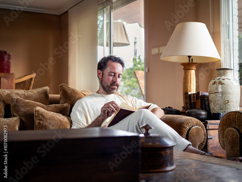 In a warmly decorated living room, a man with a beard is sitting comfortably on a sofa, engrossed in reading a book. The sunlight filters through the windows, creating a peaceful atmosphere. photo