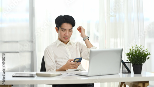Smiling young entrepreneur in white shirt sitting in office and using smartphone