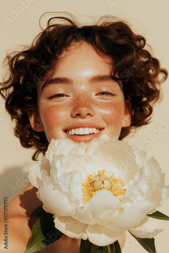 A cheerful portrait of a young woman with curly hair, smiling while holding a fresh white peony.