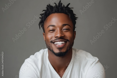 A black African man in white casual attire, smiling against a gray background, projects warmth and approachability with a charming demeanor.