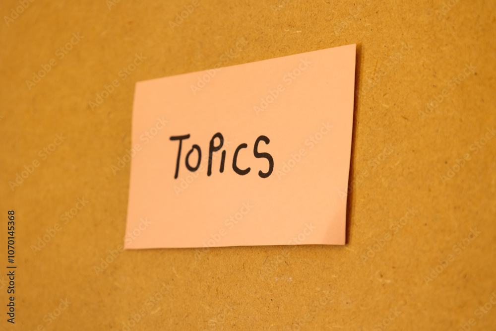 Close up of pink note stuck on a board with written message saying topics, concept of notes and topics Close up of pink note stuck on a board with written message saying topics, concept of notes and topics