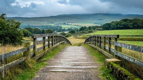 A wooden footbridge leading to a rural landscape with rolling hills and green fields under a cloudy sky.