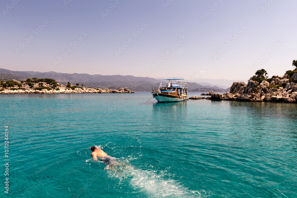 Fototapeta premium A serene image of a person swimming in the crystal-clear turquoise waters near a boat in Kaş, Turkey, surrounded by rocky islets and distant mountains
