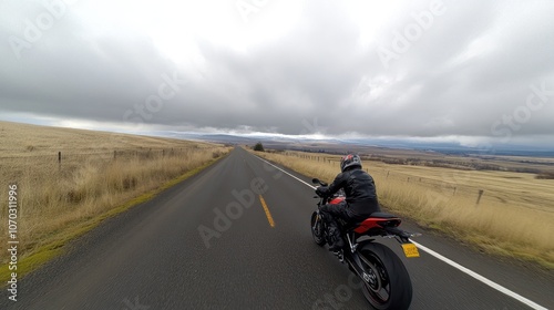 A motorcyclist rides along a wide, empty road surrounded by fields under a cloudy sky.