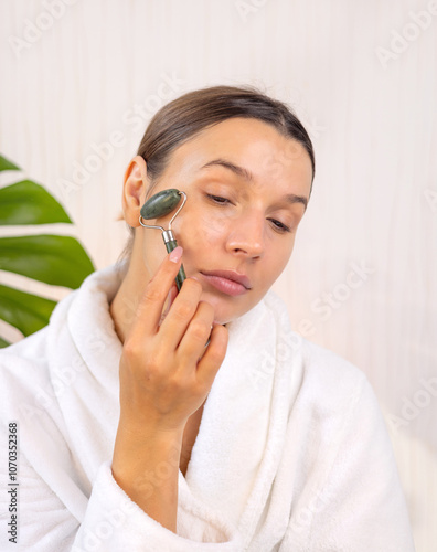 woman applying face sheet mask at home, cream or serum and massaging skin with roller.girl doing beauty routine at home. double side mirror and cream jar on table,monstera plant leaf in background.