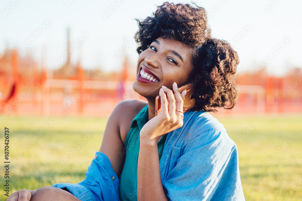 Fototapeta premium Portrait of cheerful dark skinned woman with curly hair smiling during conversation on mobile phone outdoors, attractive african american hipster girl making call sitting on green grass in park