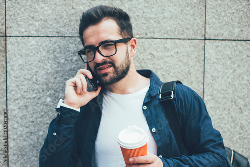 Pensive caucasian hipster guy in eyewear havin smartphone conversation standing near wall promotional background , handsome male student casual dressed making call in roaming on coffee break photo