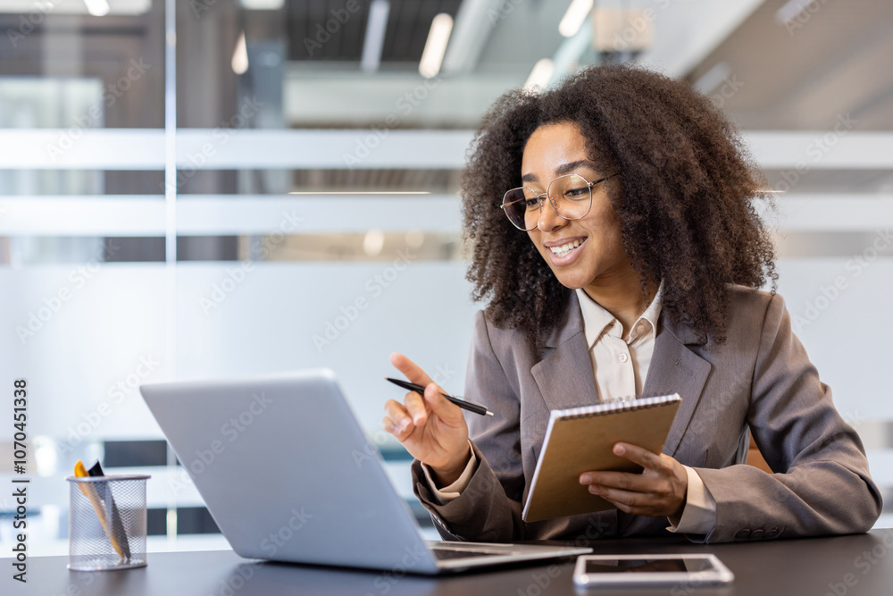 African American young woman sitting at a desk indoors, holding a notebook and a pen, talking and explaining on a video call of a laptop African American young woman sitting at a desk indoors, holding a notebook and a pen, talking and explaining on a video call of a laptop