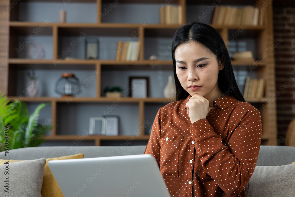 Thoughtful Asian woman using laptop at home, focusing intently on work or online learning. Comfortable setting enhances sense of concentration and productivity, surrounded by modern bookshelf. Thoughtful Asian woman using laptop at home, focusing intently on work or online learning. Comfortable setting enhances sense of concentration and productivity, surrounded by modern bookshelf.