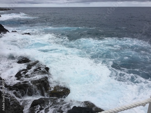 Powerful ocean waves crash against the rugged rocky shore, creating a dramatic seascape under a moody, overcast sky. The white foam adds dynamic contrast to the dark rocks.
