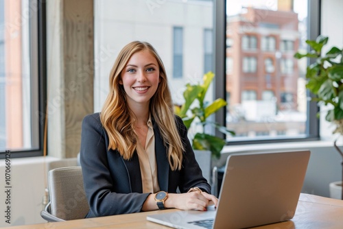 A young woman smiles while sitting at a desk in a contemporary office. She is engaged in her work on a laptop, with large windows showcasing an urban landscape behind her