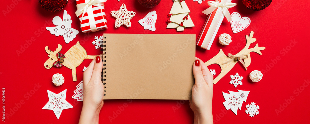 Naklejka premium Top view of female hand making some notes in noteebok on colorful background. New Year decorations and toys. Christmas time concept