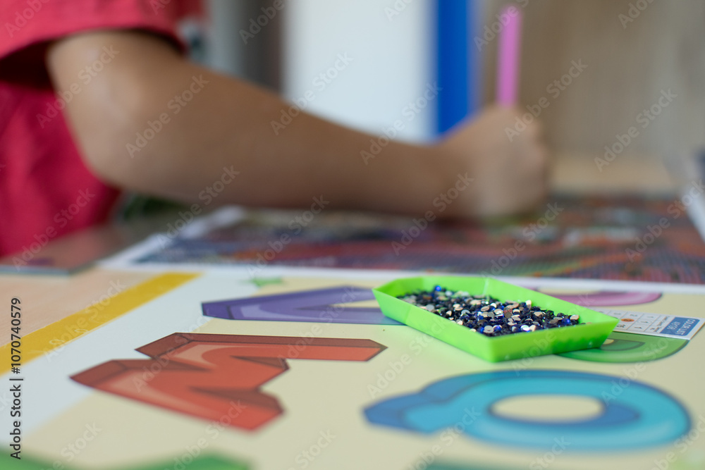 Fototapeta premium A child working on a colorful diamond painting of a lion, with alphabet learning materials and a pink stylus on a wooden desk.