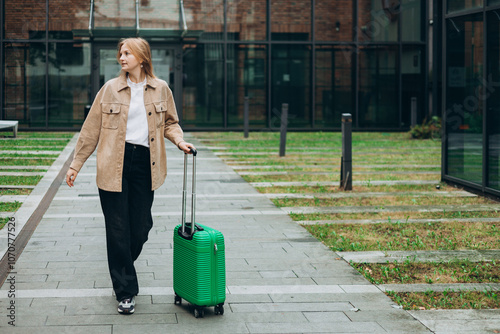 Obraz na płótnie Cheerful young woman in casual outfit walking on the city park with green luggage. 30s Woman dragging suitcase near hotel. Travel Fashion concept. Full body w salonie