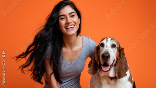 A cheerful young woman of Middle-Eastern descent smiles alongside her loyal Basset Hound against a vibrant orange backdrop.