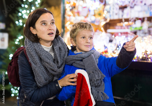 Cheerful tween boy having fun at Christmas street market with his mother, looking in amazement at festive decorations