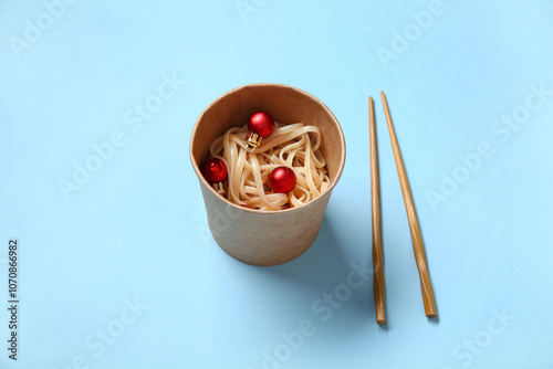 Takeaway box with chinese noodles, Christmas balls and chopsticks on color background