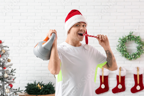 Young sporty man in Santa hat training with kettlebell and eating candy cane in living room decorated for Christmas photo