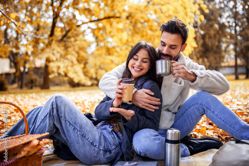 Young happy couple having picnic in a grass in a colorful autumn park. They drink coffee from thermos.