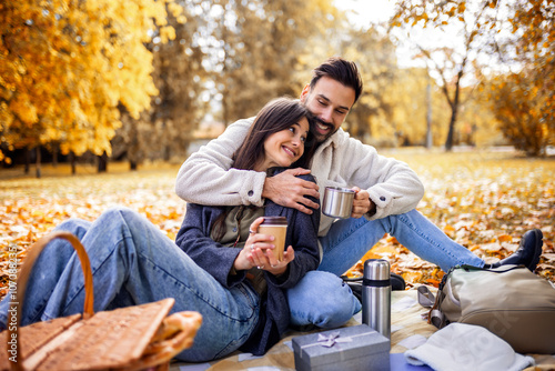 Young happy couple having picnic in a grass in a colorful autumn park. They drink coffee from thermos.