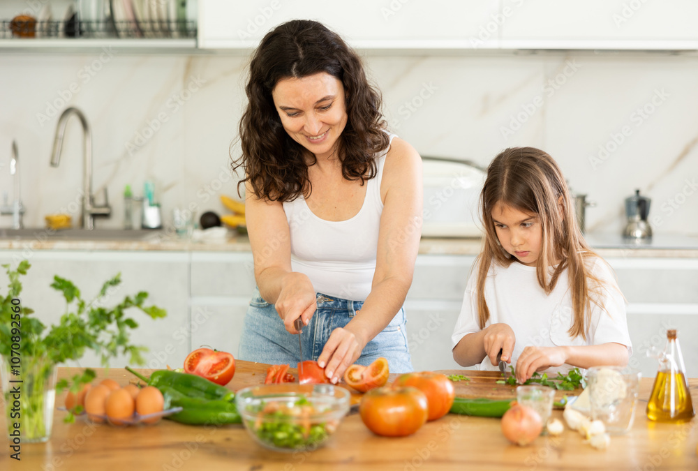 In kitchen, mom and daughter are cutting fresh vegetables to make salad. Groom explains to girl rules of cooking, peculiarities of using seasonings. Mother passing family recipe to her child. In kitchen, mom and daughter are cutting fresh vegetables to make salad. Groom explains to girl rules of cooking, peculiarities of using seasonings. Mother passing family recipe to her child.