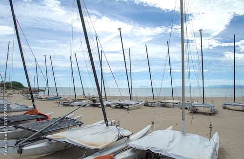 Two rows of catamarans with masts docked on the beach with sea and cloudy sky. 
 photo