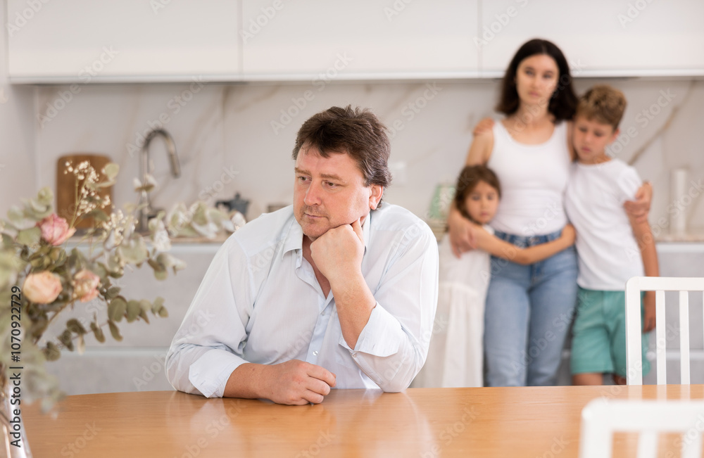 Thoughtful father is sitting in kitchen and thinking about ways to solve difficult financial situation. Wife and children stand behind dad and are afraid to ask about current problem Thoughtful father is sitting in kitchen and thinking about ways to solve difficult financial situation. Wife and children stand behind dad and are afraid to ask about current problem