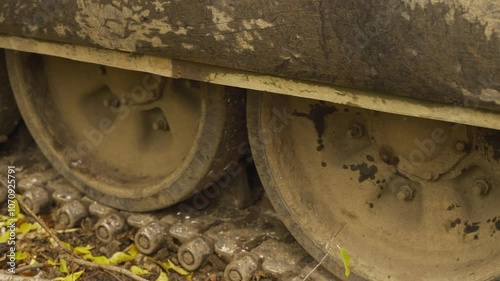 Close-up of muddy tank wheels and tracks, partially covered in dry leaves and twigs. The wheels show signs of use with mud caked on the rims	

