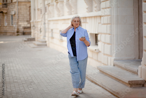 A woman aged 75-80 walks through the street of the city with a phone in her hands. An elderly madam is wearing jeans, glasses and a shirt