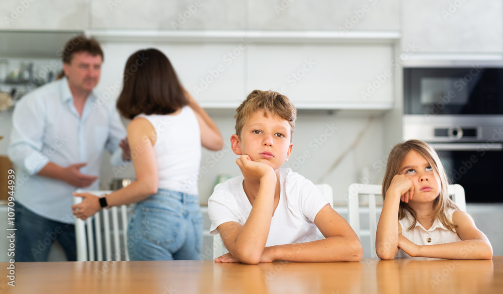 Two children sitting at the table sadly and their mother and father bickering behind in the kitchen
