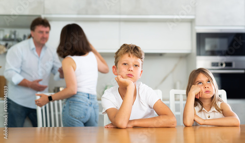 Two children sitting at the table sadly and their mother and father bickering behind in the kitchen