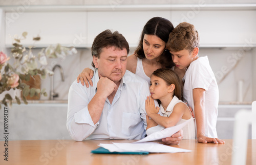Wistful father looking through papers sitting in the kitchen, his wife and children hugging him with compassion