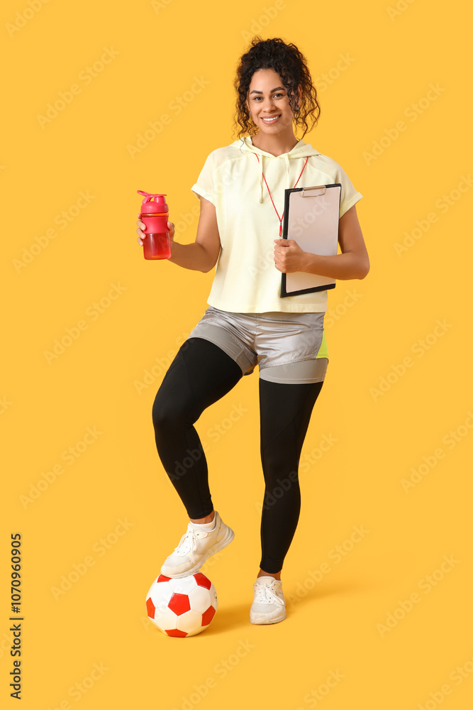 Naklejka premium Female African-American soccer trainer with ball, clipboard and water bottle on yellow background