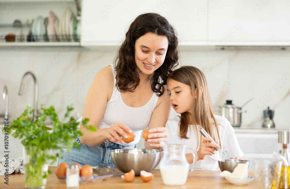 Mom teaches her daughter how to make dough, breaks eggs into bowl. Woman explains to her daughter successful recipe and rules for making dough for dessert. Homemade food and little helper. Mom teaches her daughter how to make dough, breaks eggs into bowl. Woman explains to her daughter successful recipe and rules for making dough for dessert. Homemade food and little helper.