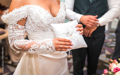 A bride in an elegant, lace-embellished wedding dress holds a white ring pillow adorned with pearls and ribbon, symbolizing love and commitment. The groom stands beside her, adding to the special mome photo