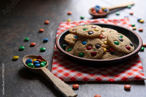 Group of red, blue, green and yellow vanilla chocolate chip cookies on a round black plate on a red napkin with white checks, surrounded by chocolate chips