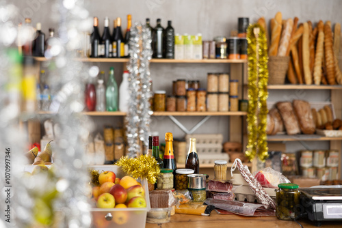 Variety of goods on counter, vacuumed meat delicacies and cheeses, fresh apples. Shopping hall of grocery minimarket is decorated with tinsel for New Year. In background there are shelves with bread photo