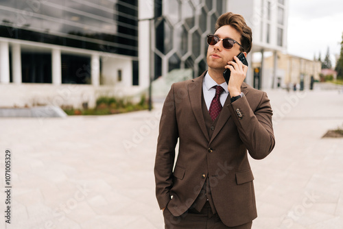 Elegant businessman in brown suit and stylish sunglasses conversing on mobile phone outside city office building, showcasing modern corporate lifestyle. Concept of business communication.