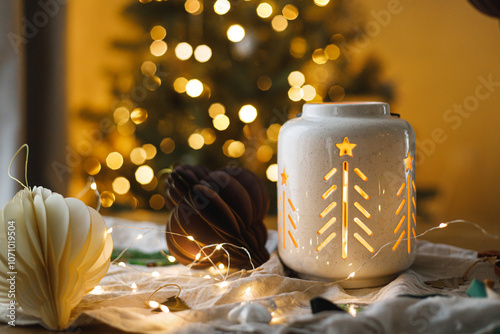 Merry Christmas and Happy holidays! Stylish glowing lantern, modern paper ornaments on table against golden christmas tree lights. Atmospheric christmas eve, holiday magical time