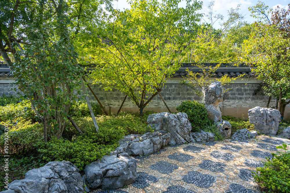 Stone path adorned with decorative pebbles is lined with ornamental rock formations in a traditional Chinese garden.Trees, shrubs,and groundcover against a boundary wall.Asian landscaping aesthetic Stone path adorned with decorative pebbles is lined with ornamental rock formations in a traditional Chinese garden.Trees, shrubs,and groundcover against a boundary wall.Asian landscaping aesthetic