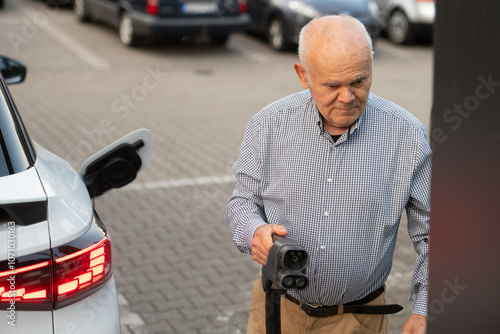 Senior Man Holding EV Charging Plug Near Electric Car in Parking Lot