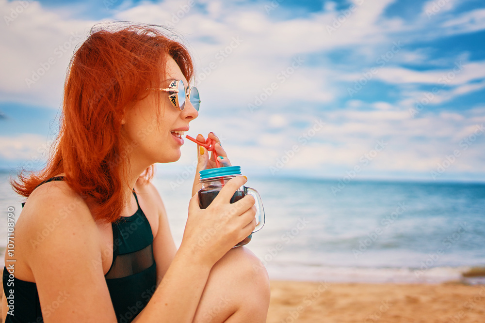 Calm lady in a swimsuit is sitting on beach with a cocktail and look away. Tropical alcohol beverage. Summer day with cloudy sky. Red-haired woman with sunglasses on vacation. Holidays at the sea. Calm lady in a swimsuit is sitting on beach with a cocktail and look away. Tropical alcohol beverage. Summer day with cloudy sky. Red-haired woman with sunglasses on vacation. Holidays at the sea.